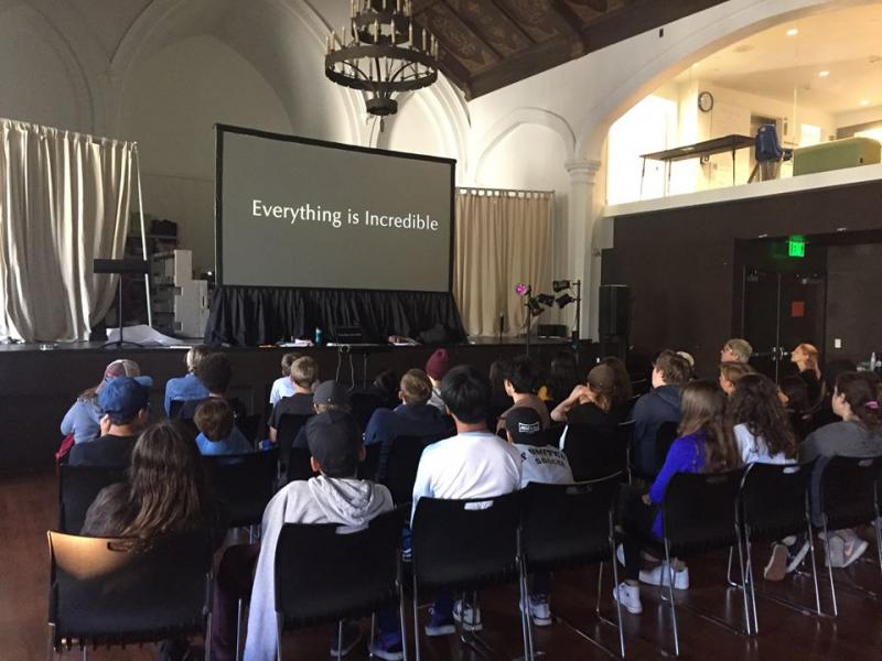 A seated group of children, youths, and adults are watching a screen with the words "Everything is Incredible" displayed.